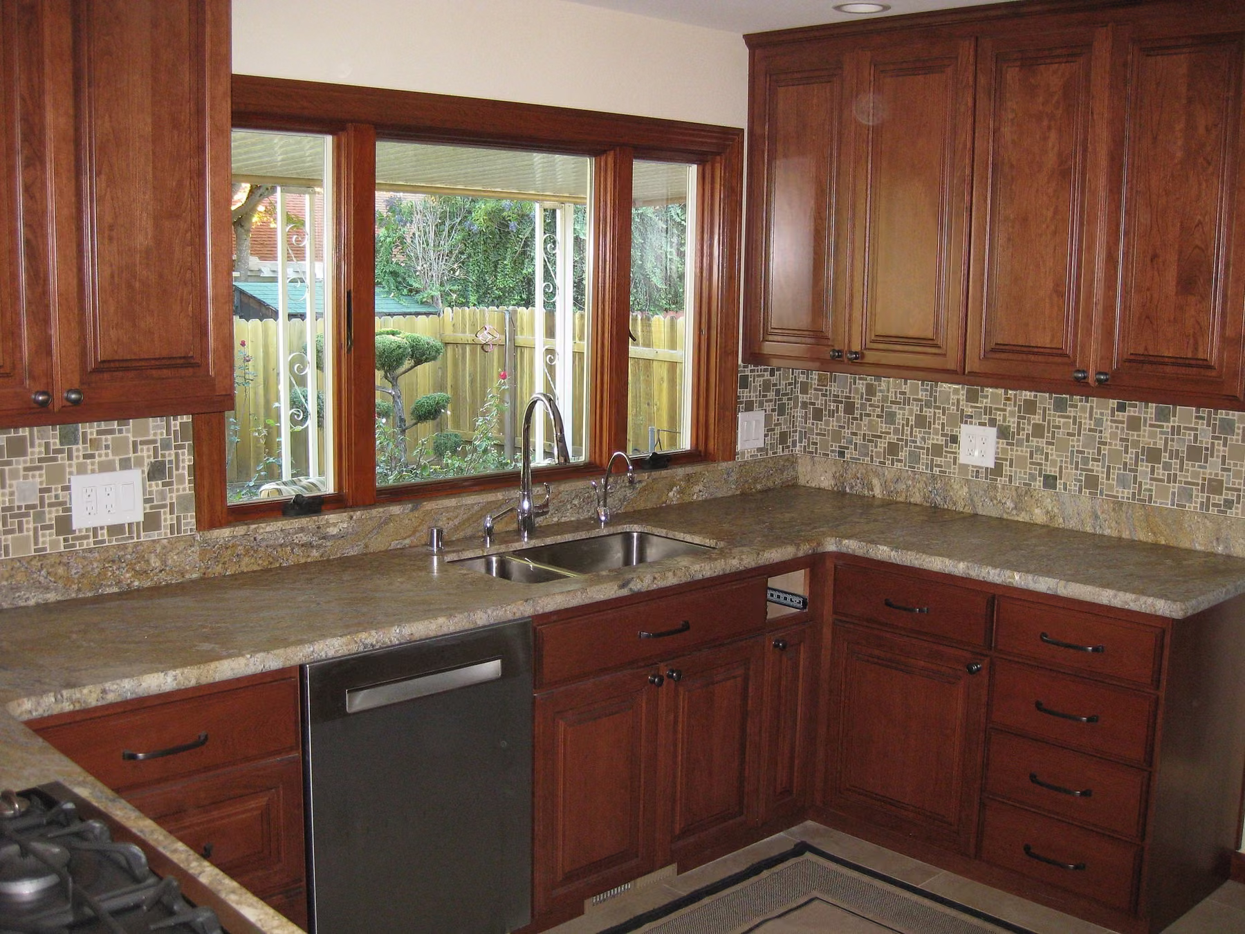 Corner kitchen with dark cherry cabinets, a mosaic tile backsplash, stainless dishwasher and a garden window over the sink