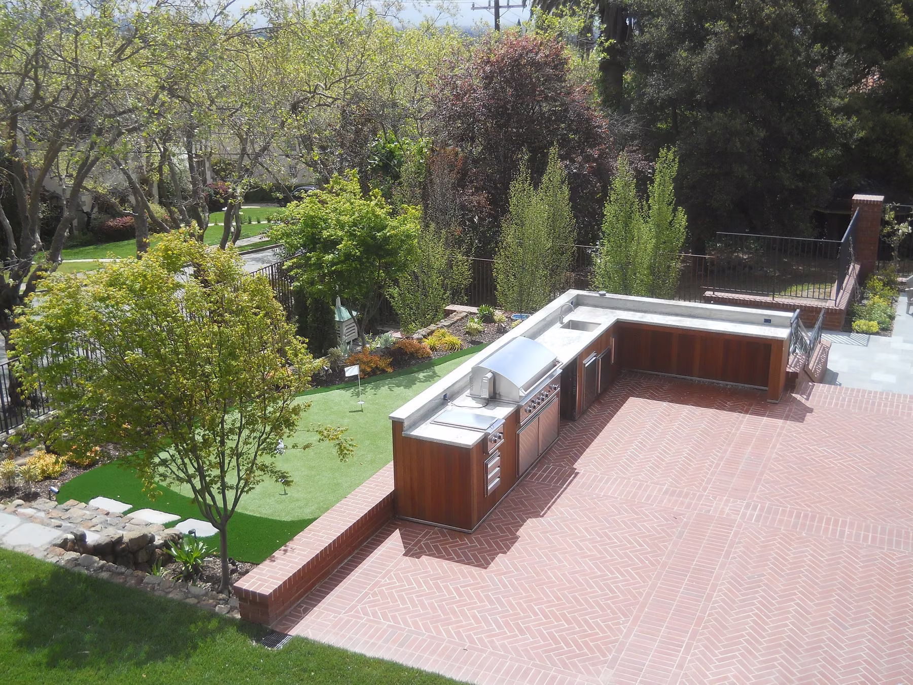 Outdoor kitchen and bar built into a raised brick terrace, seen from above with lawn and mature trees beyond